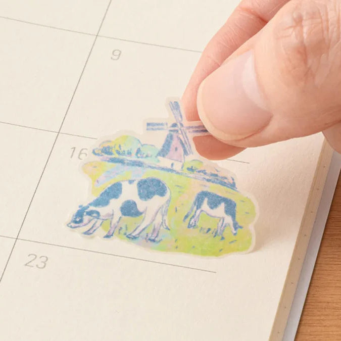 Hand placing a decorative sticker with cows and a windmill on a calendar page.