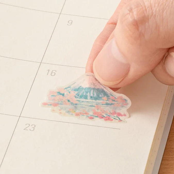 Hand placing a small, colorful sticker of Mount Fuji on a calendar page